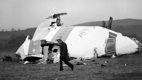 A police officer walks past the wrecked nose section of Pan Am flight 103 in a field at Lockerbie after the plane was blown apart by a terrorist bomb. The picture is in black and white. The plane is light coloured with writing on the side and the police officers is wearing all dark colours.