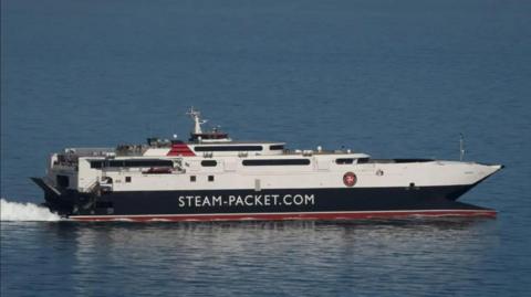The Manannan at sea on a calm sunny day, moving from the left to right. It has a white top and dark hull with a line of red above the sea line. It says Steam-Packet.com on the side in large capital letters.