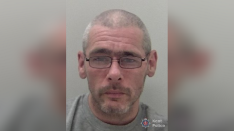 A police mug shot of a man with a shaved head and glasses. 