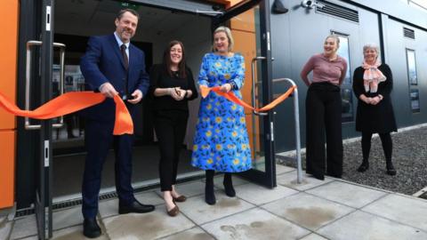 Councillor Gary Allen with short brown hair, blue suit, white shirt and maroon tie holding a cut orange ribbon with volunteer Natalie Whittaker who has long brown hair, black top, black trousers, brown shoes and is holding a pair of scissors. Councillor Pamela Hargreaves has short blonde hair with a blue patterned dress, black boots and is holding a cut orange ribbon. Leader of Hartlepool Borough Council, Danielle Connolly has blonde hair tied back with a peach coloured top and black trousers. Start Head of Service, Sally Harris and Clinical Lead has short grey hair, glasses, a black dress, black leggings and black shoes. They are stood outside the entrance to an orange and dark grey cladded building officially opening it by cutting the ribbon.