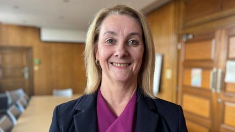 A woman with shoulder length blonde hair is smiling for a head and shoulders shot and wearing a black blazer and a purple top. The background is blurred but you can make out a conference room with wooden walls and doors.