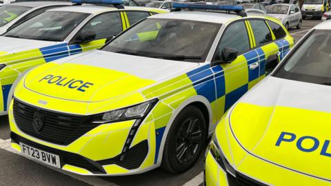 Yellow Peugeot 308 estate police car in a car park, with fluorescent blue and yellow stickers and blue light panel on the roof.
