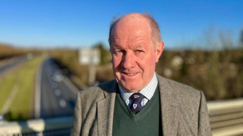 Tim Passmore with very short grey hair, smiling at camera, wearing a grey jacket, green sweater and white shirt with blue tie which has a white loco. He is standing on a road bridge - cars are visible on the road behind him.