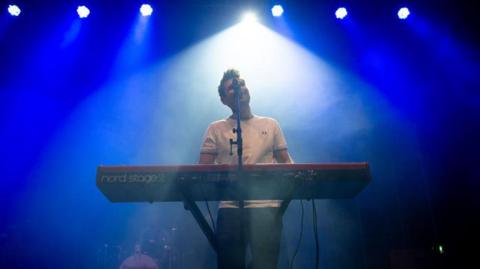 Roy Stride of Scouting For Girls singing and playing a keyboard on stage with blue and white lighting in the background