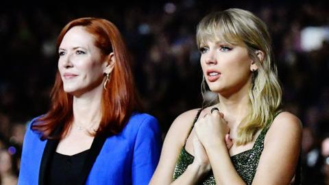 Tree Paine and Taylor Swift, standing together at an awards ceremony. Tree is wearing a blue blazer, and Taylor is wearing a sparkly dress. Taylor looks worried, while Tree has a straight face.