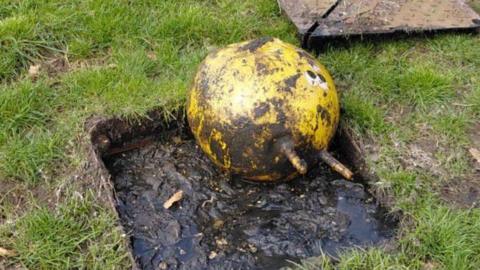 Open roadside utility hole filled with muddy water, containing a dirty yellow space hopper with two protruding handles, with the metal cover removed and lying on the grass nearby.