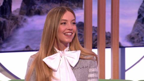 Sydney Sweeney smiles in The One Show studio, wearing a large white bowtie and a grey cardigan. She has long brown hair