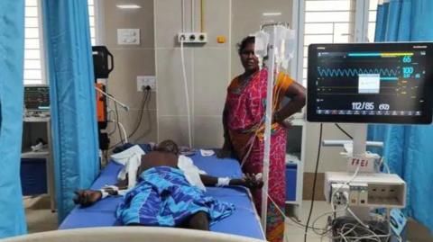 A victim lies in bed in hospital as a family member watches over him