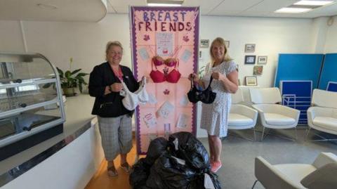 Two women stand in a canteen to either side of a bright pink banner with two pink bras attached to it. Writing on the banner reads: "BREAST FRIENDS" in capital letters. The woman on the left has blonde hair tied in a ponytail and is wearing a black jacket, checked grey trousers, brown sandals and a pink lanyard. She is holding a white bra. The woman on the right has shoulder-length blonde hair and is wearing a white patterned dress and pink trainers. She is holding a black bra.