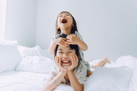 Two siblings on a white bed in a white bedroom. One lies on top of the other's back, both are smiling widely