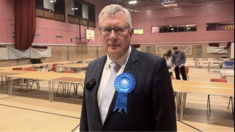 Roger Hirst with grey hair and wearing a navy suit with a white shirt and tie standing an leisure centre hall.