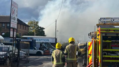 Two firefighters stand beside a fire engine. They are looking towards a car repair compound with a cloud of white smoke behind it.