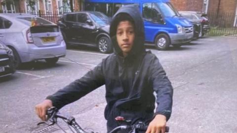 A 16-year-old boy poses on his bike, he is wearing a black jacket with the hood up. Cars and a blue van are parked behind him.