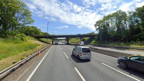 Two bridges can be seen over a dual carriageway. Trees and greenery can be seen on either side.