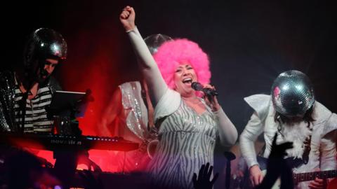 A group on stage. They are wearing 70s attire, including glitterball helmets. The woman in the middle has pink hair, probably a wig.