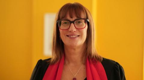 Julieanne Mitchell has reddish brown straight hair that falls just below her shoulders and a fringe above her red and dark brown-framed large glasses. She is looking directly into the camera and smiling, while wearing a red scarf and a black jacket, with a necklace that has a red heart pendant. The room behind her is painted bright yellow.