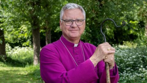 A man in his 60s with grey hair and glasses is wearing purple robes, a white collar and holding a hooked staff.