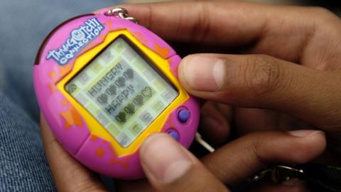 A close up shot of a Tamigotchi being held in the hands of a young girl. The device is pink and yellow, and says the works 'Tamigotchi connection' on the front, alongside 'hungry' and 'happy' on the device's display. Picture taken in Canada in 2014