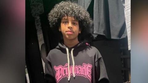 Mohanad Abdullaahi Goobe is photographed with black curly hair. He is pictured wearing a dark hoodie with bright pink writing on it.