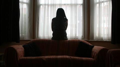 A young woman suffering from domestic violence stands alone in the bay window of her home. 