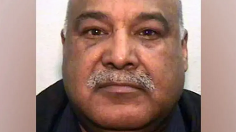 Police custody image of Shabir Ahmed who has a grey receding hair and moustache. He is wearing a black top.