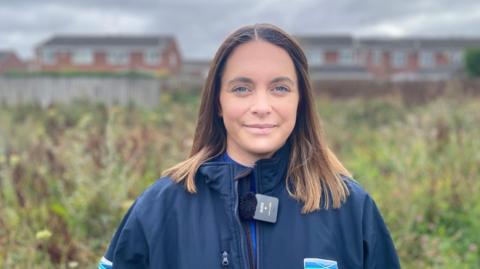 A young woman with blue eyes and shoulder-length light brown hair smiles at the camera. She is wearing a blue waterproof jacket and standing in front of an area of wild flowers and grasses with modern housing visible behind a wooden fence in the background.