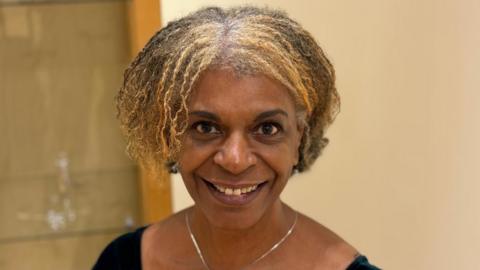 Norma Banton has short brown and blonde hair, and is smiling at the camera. She is wearing a black top and silver necklace 