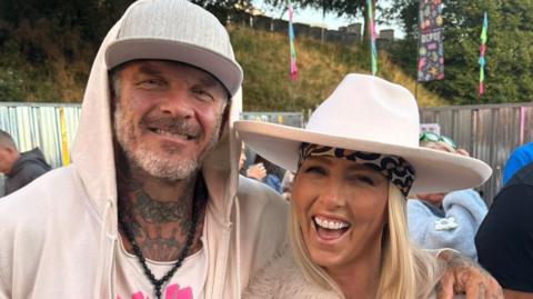 Photograph of Mathew Pritchard and his partner Ciara Dunne. Mathew wears a cream zip up hoodie, a white vest shirt and a black beaded necklace. He wears a grey flat cap hat, has a silver beard and blue eyes and smiles at the camera. His wife wears a patterned scarf under a white wide brimmed hat, and a fur jacket.