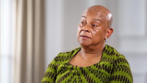Doreen Lawrence wears a green dress with black patterning, and is sitting in a large formal room