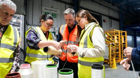 Four people wearing high-viz vests examine yarn from which parachute material is made, which is on big spools on a table. To the right a pair of hands can be seen while in the background is panelling and yellow shelves as well as a large door. 