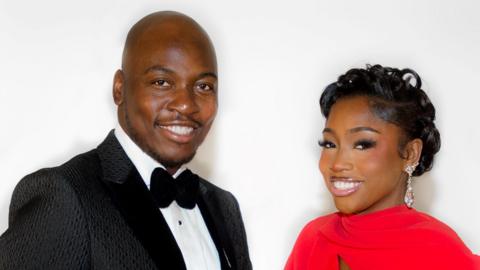 Eddie Kadi and Indiyah Polack in a promotional shot for the Mobo Awards. Eddie is dressed in a black tux with a black bow tie and holds a Mobo trophy. Indiyah wears a high-necked red dress, her hair curled on top of her head. 