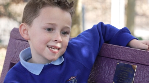 Teddy, is seen smiling as he sits on a purple bench made from recycled plastic tubs. He is wearing a light blue shirt under a blue top