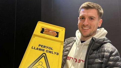 Dion's friend Tom poses with the sign. Perygl llawr glwyb  which translates to wet floor hazard can be seen at the top of the sign.