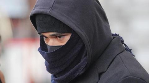 Close up of Adeel Khan from the shoulders up. His face is largely obscured apart from his eyes and eyebrows by a black hat and face covering. He is also wearing a black scarf and black hoodie over his head.