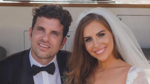 Theo Schooling and his wife on their wedding day. He has dark hair and is wearing a black suit with a bow tie, and she has brown hair and is wearing a veil.