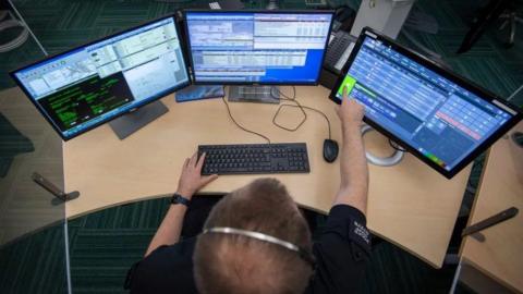 A photo of a police dispatch officer from above. There are three screens on the desk in front of the uniformed man as well as a keyboard and mouse. The man is wearing a headset and is pointing to a green box at the bottom of the right-most screen. The screen show an indistinct array of boxes full of written information and data.