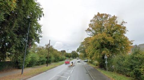 A tree-lined 30mph suburban road, flanked by footpaths and featuring a traffic island. 