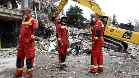 Three emergency workers in red uniforms stand near a yellow digger as it removes rubble
