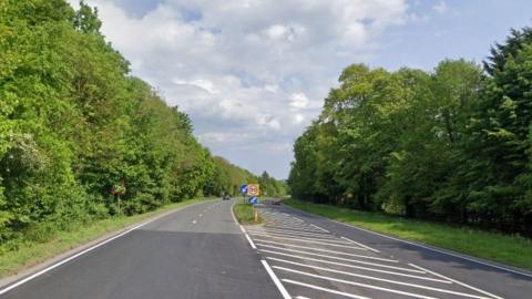 A section of the road, where a single carriageway turns into a dual carriageway. The road is lined with dense trees and bushes. 