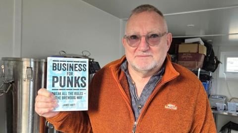 A man with glasses wearing an orange fleece holding a blue book "Business for Punks" by Brewdog founder James Watt