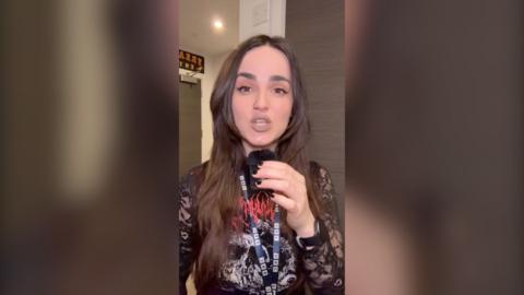 Woman with long dark brown hair holding a small microphone, looking straight at the camera