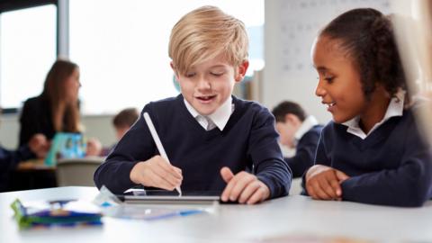 A stock image shows a classroom scene with children in school uniforms seated at a table, with one child using a digital tablet and stylus while another looks on. Other pupils and learning materials are visible in the background.