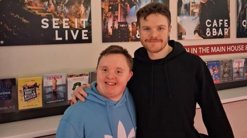 A young man with Down's syndrome, wearing a light blue hoodie, stands next to a taller man with brown hair and a short beard, wearing a black hoodie. They stand in front of a display of leaflets and posters for theatre performances.