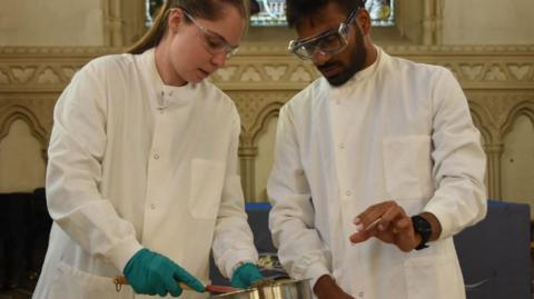 Jennifer Palmer with hair pulled back wearing blue gloves and Satish Viswanathan, with dark hair and beard, both wearing white lab coats and looking down at bowl