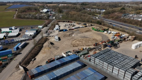 A photo taken from a drone, showing a stretch of industrial land with cars and lorries parked on it. There's a road on the left hand side and a motorway on the right.