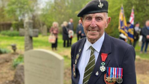 Ken Cooke wears a military uniform and stands next to a headstone
