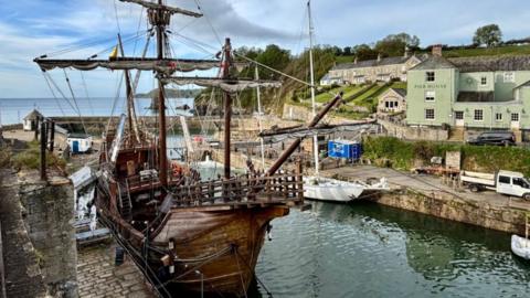 A view of Charlestown harbour. It has two stone walls forming a pincer. There is a small white watchtower building at the far end on the left. On the left in the foreground is a moored old-style wooden sailing ship. A more modern yacht is ashore on the right. On the upper right of the picture is the Pier House pub, painted in a matt light pea green.