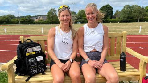 Two women sitting in running outfits on a bench with a running track behind them.