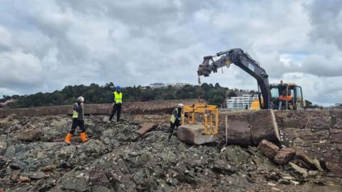 A group of workers and a crane working on a damaged seawall. The crane is lifting a large piece of stone wall with the workers helping with the works. In front of the wall is rocks covered in seaweed and shells. 