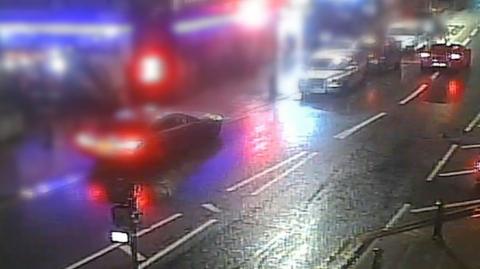 CCTV still showing cars on a road with a blurred image of individuals confronting each other outside a bar.